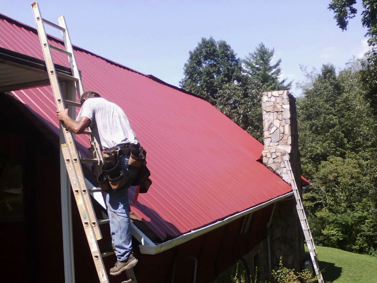 Roofer on a ladder beside a red metal roof and stone chimney — Soffit & Fascia Repair by Roofing Brothers in Justin